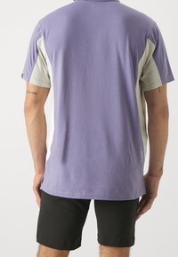 Chemise légère à manches courtes avec un corps lavande et des panneaux latéraux texturés gris clair, mettant en avant un discret logo au bas.