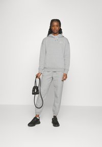 Donna che indossa una felpa grigia di Under Armour e pantaloni jogger, che tiene in mano una corda elastica nera, in piedi contro uno sfondo bianco semplice.