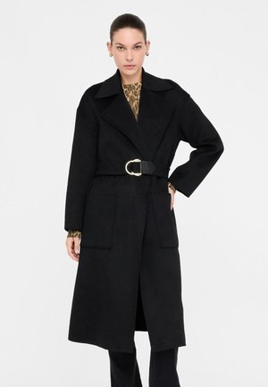 Cappotto trench in lana nero con un ampio colletto, vita accentuata da una cintura, grandi tasche frontali e silhouette dritta, indossato sopra una maglietta con stampa leopardata.