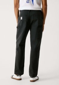 Schwarze Cargo-Hose aus strapazierfähigem Material, mit aufgesetzten Taschen, lockerer Passform und einem Logolabel auf der Rückseite. Ergänzt durch weiße Sneaker.