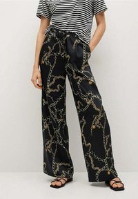 Persoon draagt een zwarte wijde broek met een gouden kettingpatroon, een gestreepte zwart-witte T-shirt met korte mouwen, en zwarte sandaaltjes met bandjes.