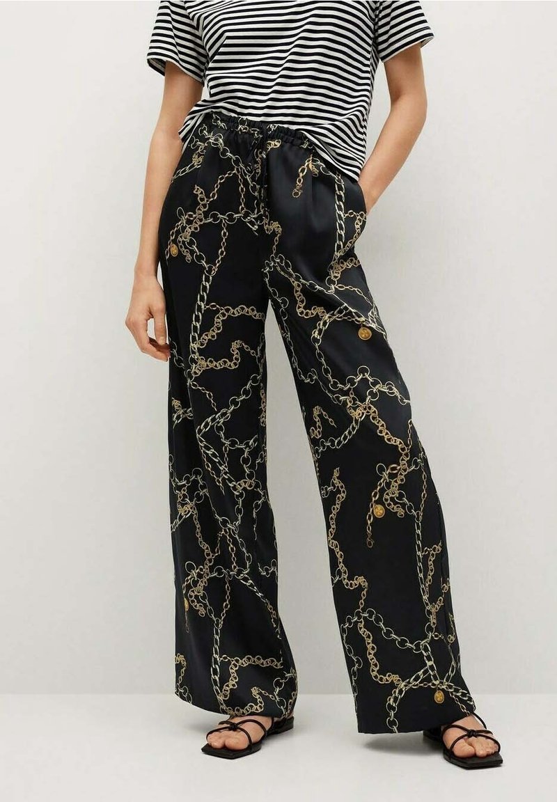 Persoon draagt een zwarte wijde broek met een gouden kettingpatroon, een gestreepte zwart-witte T-shirt met korte mouwen, en zwarte sandaaltjes met bandjes.