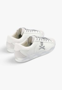 Zapatillas de cuero blanco con acentos de ante gris, que presentan cordones plateados y un diseño de calavera y huesos cruzados en el lateral. Suela texturizada.