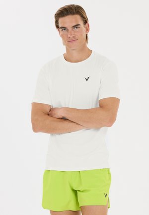 Mand med krydsede arme iført en hvid t-shirt og lysegrønne shorts, der står mod en ensfarvet hvid baggrund.