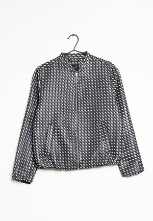 Schwarze Bomberjacke mit silbernem geometrischem Muster, Frontreißverschluss mit rundem Zipper und Seitentaschen, hängend an einem schwarzen Kleiderbügel.