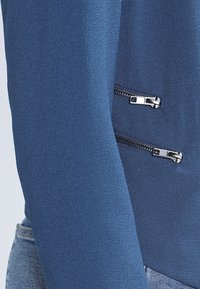 Blå långärmad topp i mjukt material, med två silvriga dragkedjor på vänster sida. Slät textur med en figurnära design.