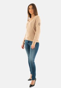 Pull beige à col en V avec des manches longues et des accents rayés gris sur le bras gauche, associé à un jean skinny bleu et des escarpins noirs à bout pointu.