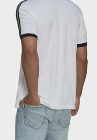 Homme portant un t-shirt blanc à manches courtes avec bordures et bandes noires sur les manches, un jean en denim bleu clair, avec une main dans la poche arrière.