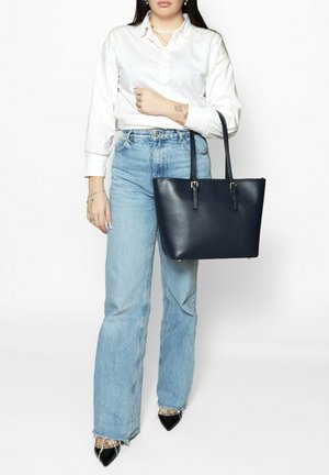 Sac en cuir bleu marine avec deux poignées et des accents métalliques, montré tenu avec une chemise de couleur claire et un jean taille haute.