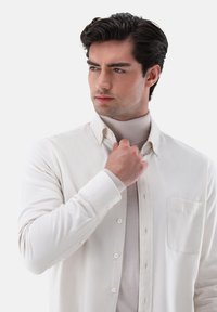 Helles, beiges Hemd mit Knöpfen und einer Brusttasche, kombiniert mit einem taillierten, hellen Pullover. Glatte Textur, klassischer Kragen und lange Ärmel.