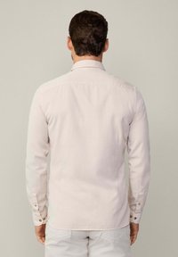 Camisa de color beige claro con mangas largas, cuello con botones y puños ajustables; textura suave y diseño sencillo visible desde la parte de atrás.