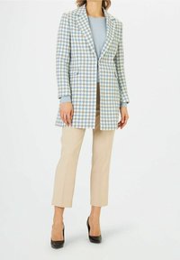Blazer à carreaux bleu et blanc, coupe ajustée, à boutonnière simple avec poches, associé à un pantalon beige et des talons noirs.