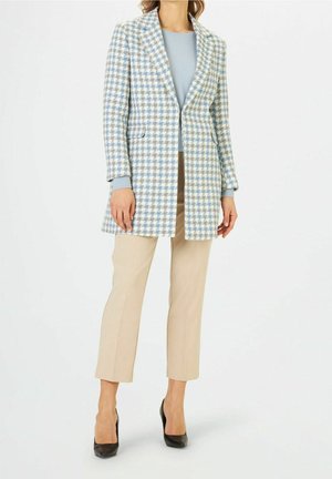 Blazer à carreaux bleu et blanc, coupe ajustée, à boutonnière simple avec poches, associé à un pantalon beige et des talons noirs.