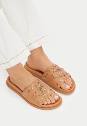 Füße, die hellbraune Ledersandalen mit Metallnieten und dekorativen Schnallendetails tragen, kombiniert mit weißen Hosen, die an den Knöcheln sichtbar sind.