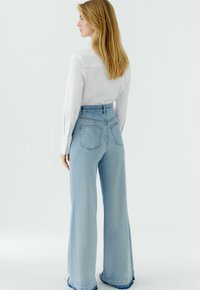 Hellblaue, hoch taillierte Jeans mit weitem Bein und ausgefranstem Saum, fünf Taschen und Gürtelschlaufen, kombiniert mit einem weißen Langarmshirt.