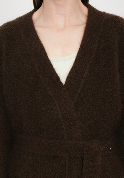 Brown knitted cardigan with a textured finish, V-neckline, and a tie belt at the waist, layered over a light-colored top.