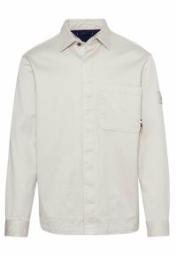 Vaaleanbeige pitkähihaisuus, jossa on napitettu etuosa, rintatasku ja hienovaraiset tikkaus yksityiskohdat. Kangas vaikuttaa sileältä ja kestävältä.