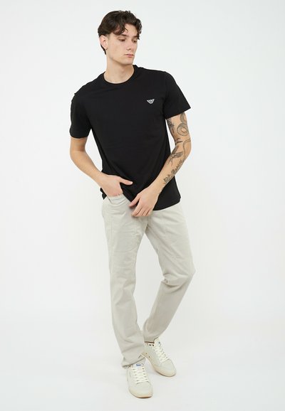 Hombre con brazo tatuado vistiendo camiseta negra, pantalones beige claro y zapatillas blancas, de pie con las manos cerca de los bolsillos frente a un fondo liso.