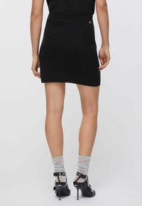 Črna mini krilo iz pletenine s prilegajočim se pasom in subtilnim zlatim poudarkom. Oblečeno s prosojnimi gležnjnimi nogavicami in čevlji z visokimi petami.