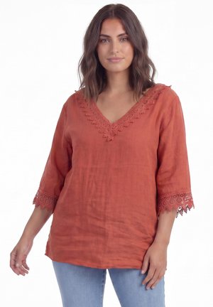 Femme aux cheveux châtain mi-longs portant un chemisier à col en V couleur rouille-orange avec une bordure en dentelle aux manches et à l'encolure, associé à un jean bleu clair.