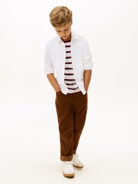 Chłopiec w białej koszuli z guzikami nosi czerwoną i białą koszulkę w paski, brązowe spodnie kordowe oraz białe trampki, patrząc w dół.