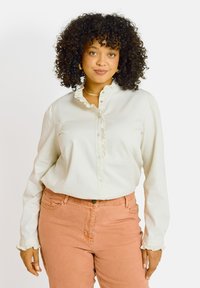 Blouse blanche à manches longues avec un col froncé et une fermeture à boutons, assortie à un jean rose pastel avec une fermeture à un seul bouton.