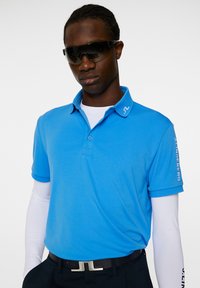 Kirkkaansininen polo-paita, jossa on pehmeä tekstuuri, lyhyet hihat ja kaulus. Logo kauluksessa ja hihassa; yhdistetty mustan vyön ja pitkien valkoisten paidan hihansuiden kanssa.