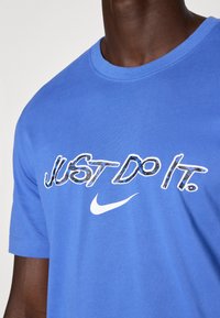 Sininen puuvillapaita, jossa on pyöreä pääntie. Etupuolella keskeisesti sijaitseva valkoisella teksti "JUST DO IT." ja valkoinen Nike-logoswosh.