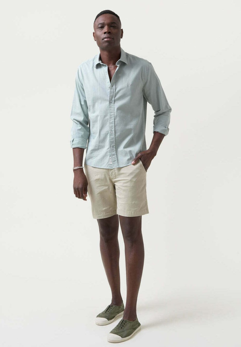Camisa de botones azul claro con mangas remangadas, combinada con pantalones cortos beige y zapatillas verdes, de pie contra un fondo neutro.