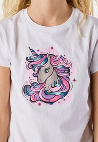 Valkoinen puuvillateeppari, jossa on unicorn-grafiikka, jossa on valuvia vaaleanpunaisia ja sinisiä hiuksia, sekä vaaleanpunaisia tähtiä ja pyöreä tausta.