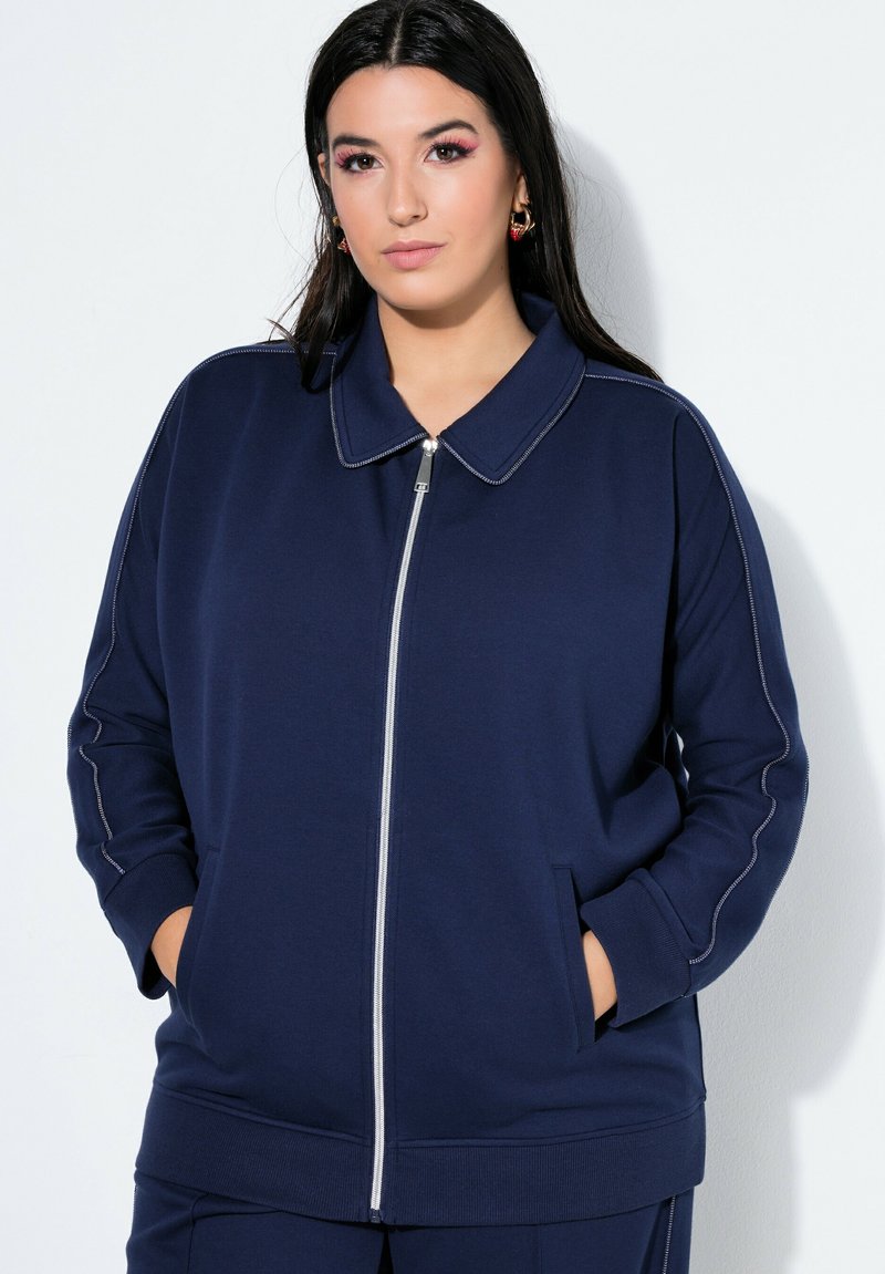 Donna che indossa una giacca blu navy con cerniera e impunture bianche, in piedi con le mani nelle tasche davanti a uno sfondo bianco.