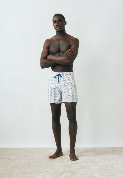 Μυώδης άνδρας στέκεται ξυπόλητος μπροστά σε λευκό τοίχο με τα χέρια σταυρωμένα, φορώντας άσπρα μαγιό με μικρά πολύχρωμα αστέρια.