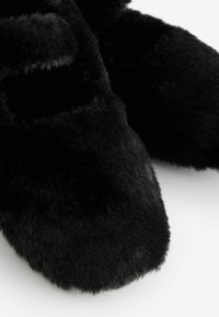 Czarne pluszowe kapcie o miękkiej, futrzastej teksturze i zaokrąglonym czubku. Posiadają dwie regulowane klamry na górze dla bezpiecznego dopasowania.