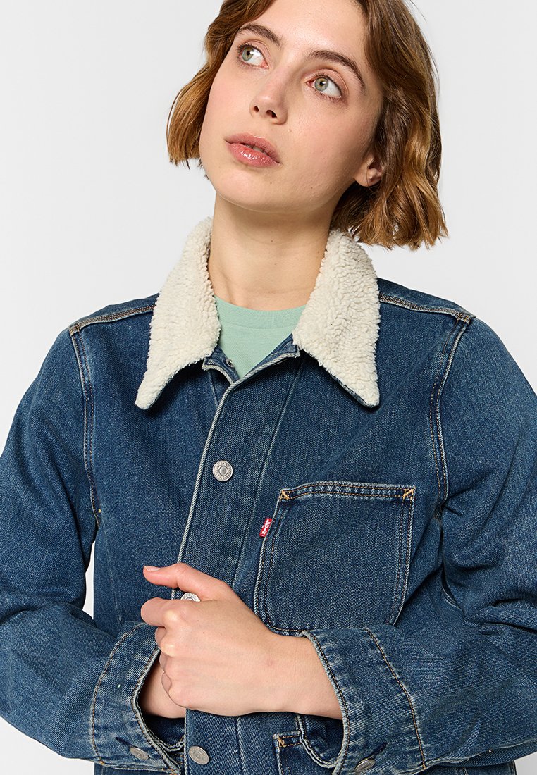 Chaqueta de mezclilla con cuello forrado de sherpa, cierre de botones, bolsillo frontal y acabado en tono azul. Costuras visibles en toda la prenda.