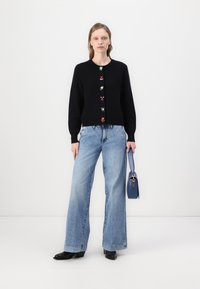 Crni kardigan s šarenim cvjetnim gumbima, uparen s svijetloplavim širokim trapericama i malom plavom torbicom. Model stoji protiv jednostavne pozadine.