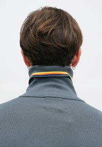 Aiz skatāmās personas silta zilā džemperī ar augstu apkakli, kas rotāta ar dzeltenām un oranžām svītrām, un ar maziem riņķiem ausīs.