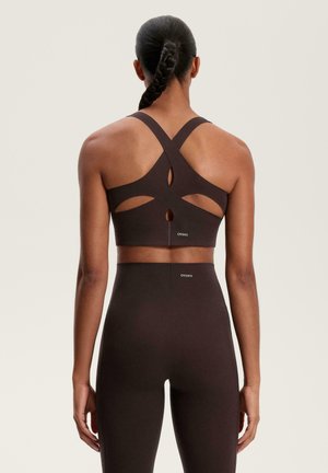 Soutien-gorge sport marrone con design a incrocio sul retro, tessuto testurizzato, dettagli con aperture e posizionamento del logo. Abbinato a leggings a vita alta coordinati.