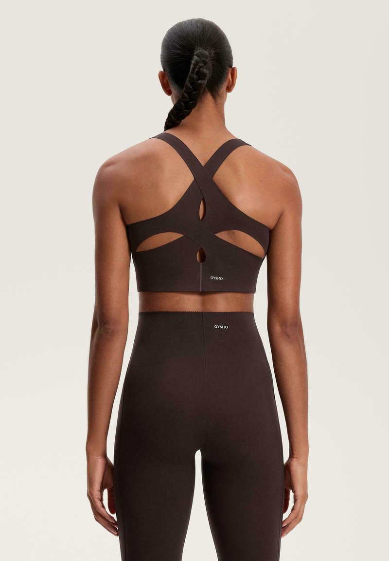 Hnědá sportovní podprsenka s křížovým designem na zádech, strukturovanou látkou, výřezovými detaily a umístěním loga. V kombinaci s odpovídajícími legínami s vysokým pasem.