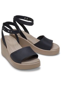 Sandali wedge neri con tomaia in gomma liscia, ampia fascia frontale e cinturini alla caviglia regolabili. Base del plateau di un leggero color beige con soletta testurizzata.