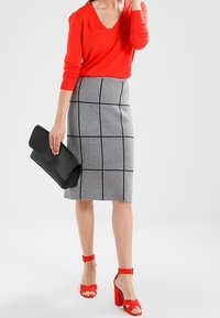 Femme portant un haut rouge à manches longues, une jupe grise à motifs de grille noire, des sandales rouges à talons hauts, et tenant une pochette noire.