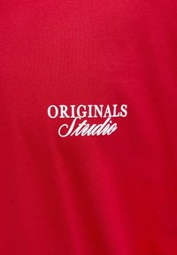 Punane puuvillane T-särk, millel on valge tikandiga tekst "ORIGINALS Studio" segas serif- ja kursiivfondides. Pehme tekstuur, liibuv disain.