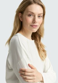 Jonge vrouw met lichtblond haar die een witte geribbelde top met lange mouwen draagt, zachtjes glimlachend met haar armen gekruist op een effen lichte achtergrond.