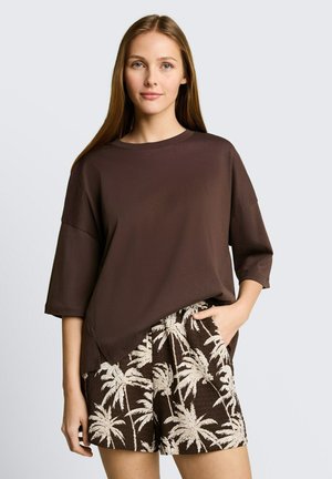 Młoda kobieta ubrana w luźny top koloru czekoladowego brązu i brązowe krótkie spodenki z białymi palmami, stoi z jedną ręką w kieszeni.
