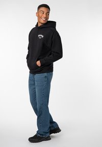 Schwarzer Kapuzenpullover mit Fronttasche, ausgestattet mit weißem Logo-Text. Getragen mit blauen Jeans und schwarzen Turnschuhen, im Seitenprofil gestylt.
