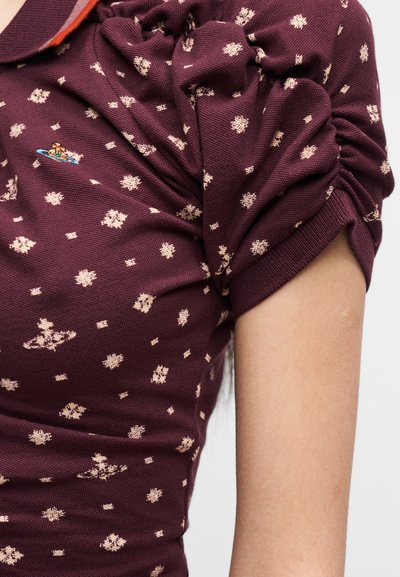 Κοντινό πλάνο ενός ατόμου που φοράει μπλούζα μαρονού χρώματος με μπεζ λουλουδάτα μοτίβα και ένα μικρό κεντημένο λογότυπο κοντά στον ώμο.