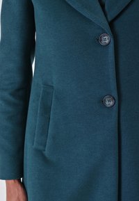 Cappotto texture verde acqua con due bottoni scuri allacciati, tasca laterale verticale e maniche lunghe su un busto e braccio di una persona.