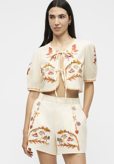 Νεαρή γυναίκα με ασορτί κρεμ crop top και σορτς με πολύχρωμη κεντητή διακόσμηση από λουλούδια και μανιτάρια, στέκεται με το ένα χέρι στη τσέπη.