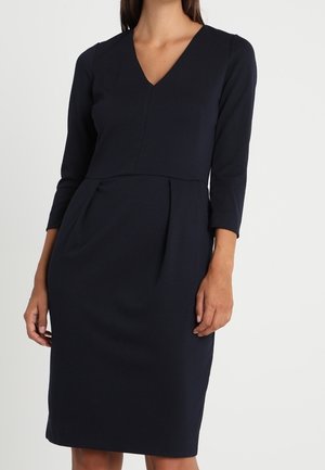 Kobieta w czarnej, dopasowanej sukience do kolan z rękawami do połowy ramienia i dekoltem w serek na jednolitym tle.