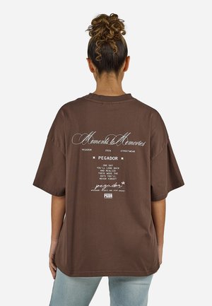 Kvinne med krøllete oppsatt hår, iført brun oversized T-skjorte med hvit trykt tekst «Moments to Memories» og lyseblå jeans, stående med ryggen til kameraet.