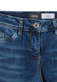 Donkerblauwe denim jeans met een gladde textuur, voorzien van gouden stiksels, een rits en een zichtbaar logo-label op de tailleband.